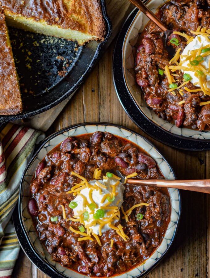 Two bowls of Beef Short Rib Chili with cornbread in a cast iron pan on the side