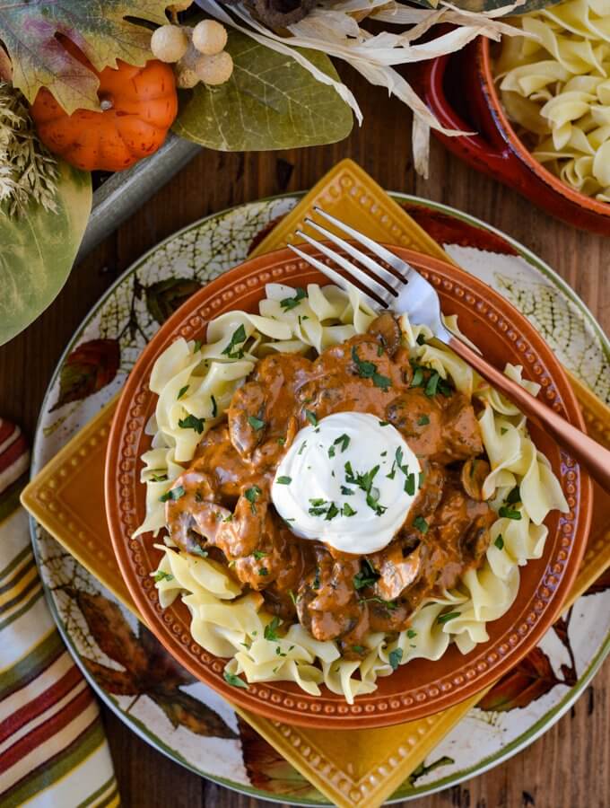 Beef Stroganoff on a bed of buttered egg noodles in fall dinnerware next to a glass of wine. There is a side dish of noodles and striped napkin.