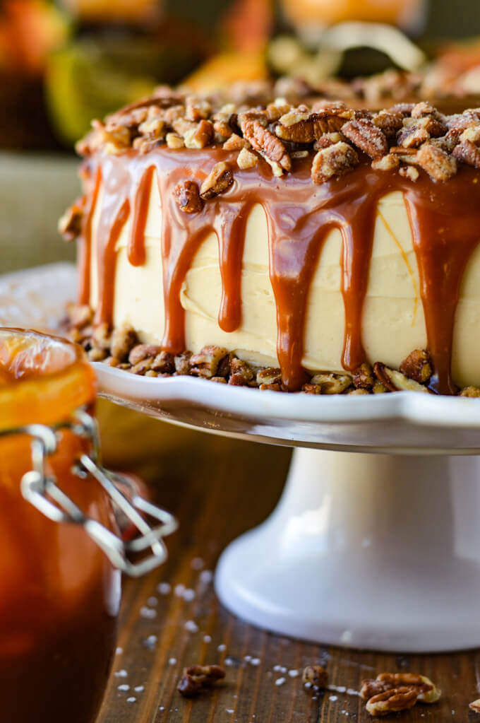 A frosted layered pumpkin cake with caramel dripping down its sides and coated on top with a layer of pecans. The cake sits on a white cake platter with a jar of caramel sauce beside it.