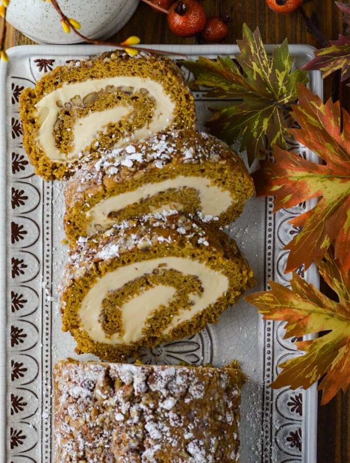 A pumpkin cake roll with three slices cut on a serving tray with fall leaves next to it.
