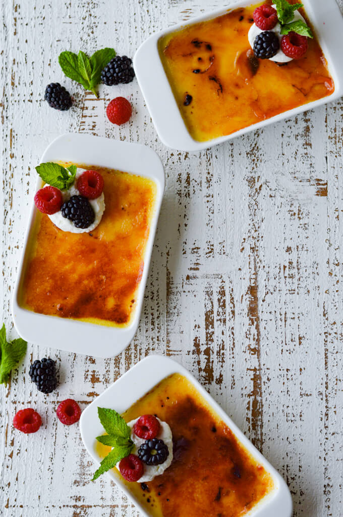 Three creme brulee with fresh fruit on a white surface with fresh berries and mint leaves scattered around.