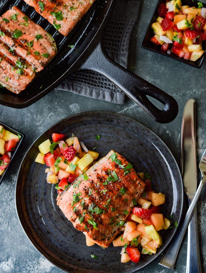 Pan Seared Salmon on a bed of fruit salsa on a charcoal color plate and a cast iron skillet next to it.