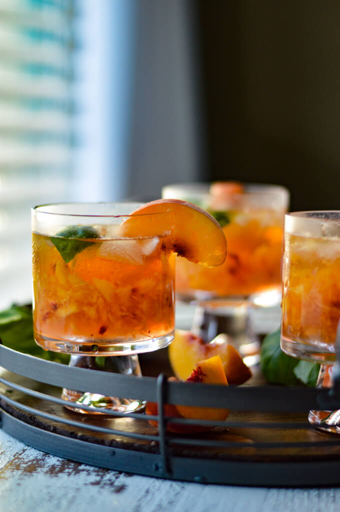 A tray of peach bourbon cocktails with fresh peach slices and fresh basil scattered around. 