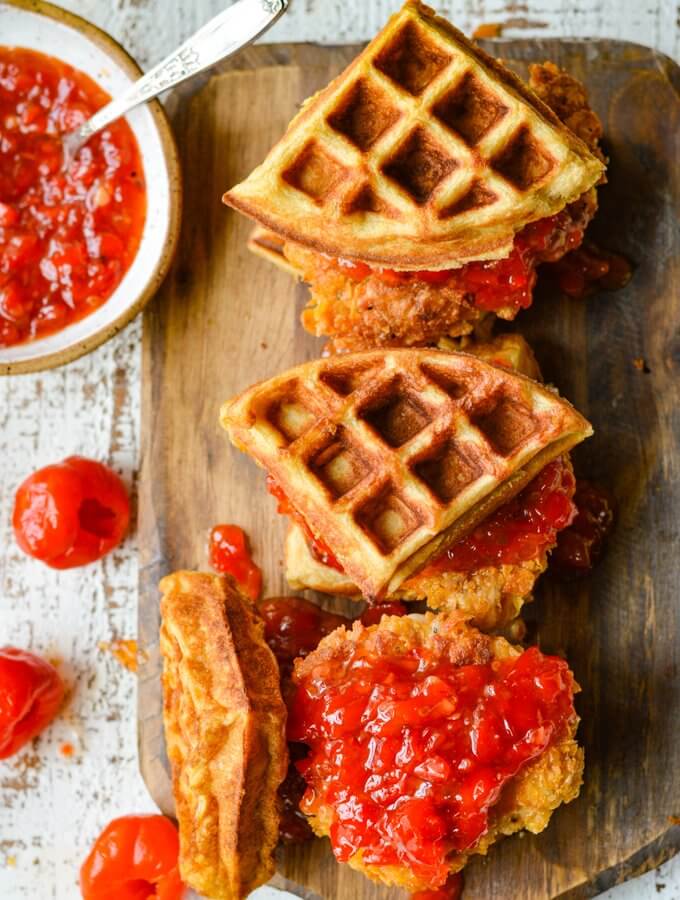 Three chicken and waffles on a board with bowl of peppadew jam and peppadew peppers scattered around.