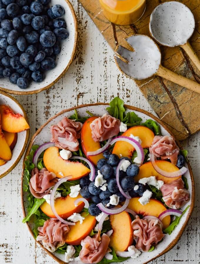 A Peach and Blueberry Arugula Salad in a bowl next to a ceramic salad fork and spoon. A bowl of blueberries and bowl of sliced peaches sit next to it.