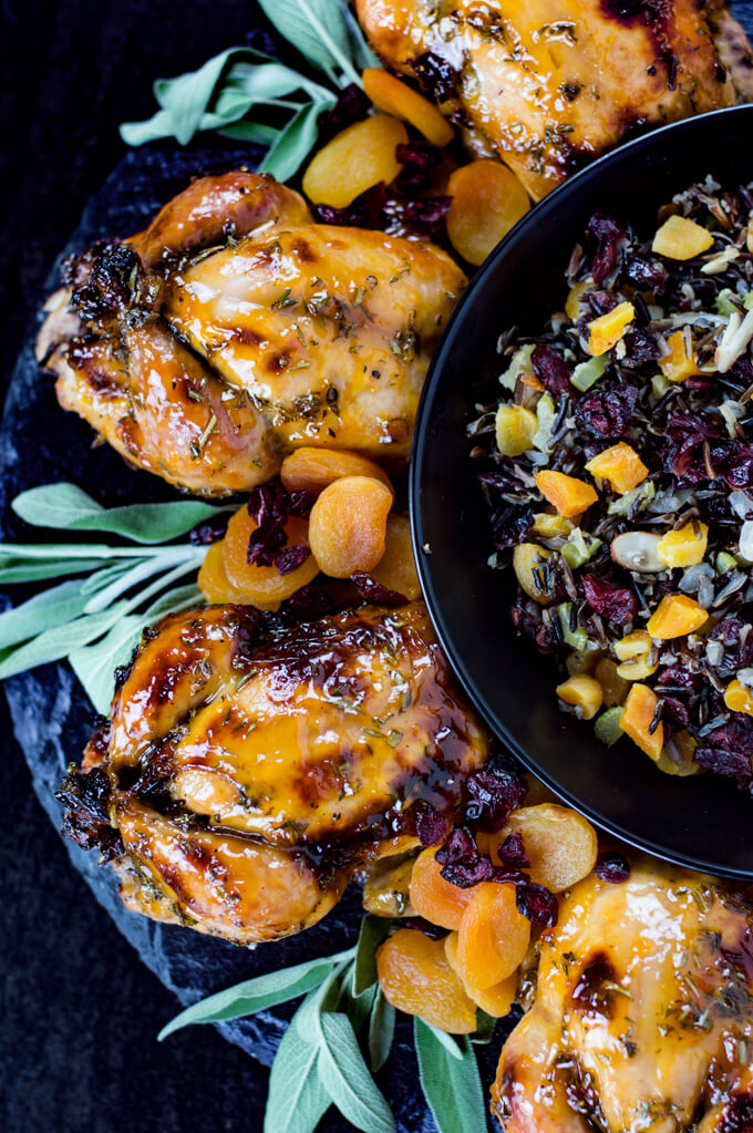 Four roasted cornish hens with an apricot glaze sit on top of a wild rice stuffing on black plates. A black bowl of wild rice stuffing sits next to it.