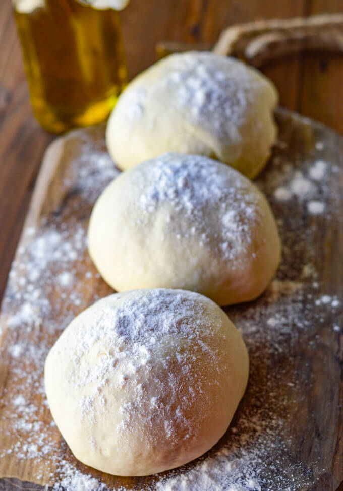 Three balls of pizza dough rest on a floured wooden board with olive oil in the background.