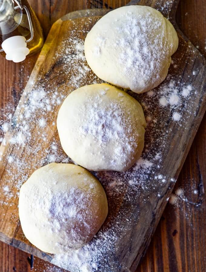 Three balls of pizza dough rest on a floured wooden board with olive oil in the background.