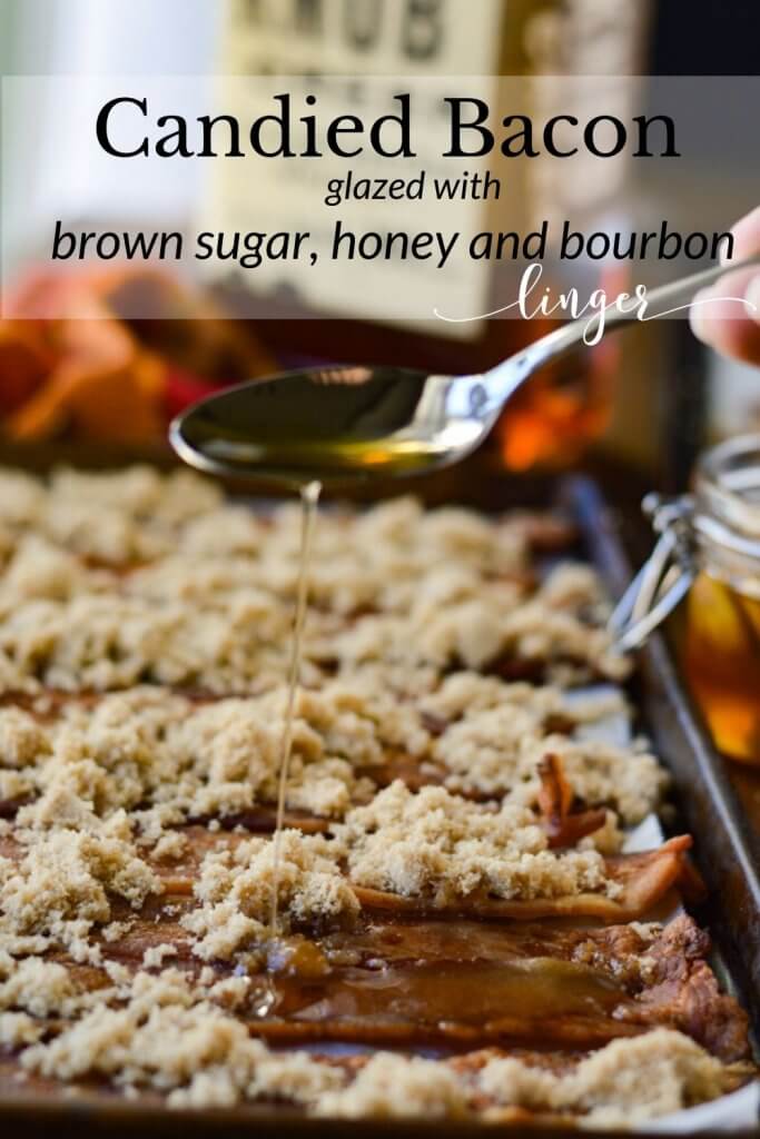 A cookie sheet with cooked bacon slices and sprinkled with brown sugar. A spoonful of honey and bourbon is being poured onto the slices. A jar of honey, and a bottle of bourbon with an orange and red napkin lays beside the pan.