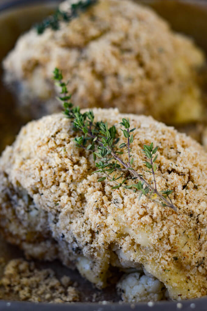 Two breaded cauliflower halves with thyme sprigs on them sit in a pan ready to go in the oven.