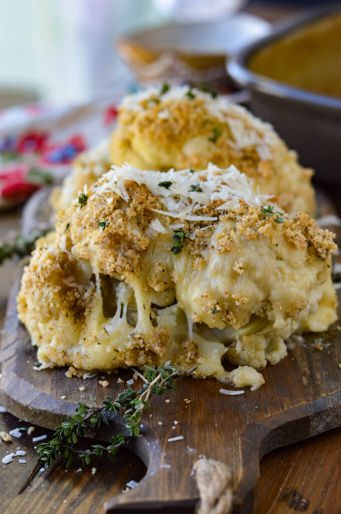 A baked breaded cheesy cauliflower half sitting on a wooden board with shredded parmesan cheese sprinkled on top and sprigs of thyme in front.