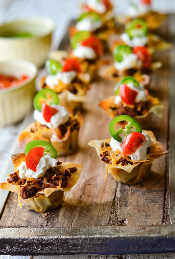 Mini Taco Wonton Cups with sour cream, cherry tomato and jalapeno slices. All of these appetizers are sitting on a wooden board with a cup of tomatoes sitting next to it.