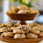A tiered wooden platter full of oatmeal raisin cookies with a bouquet of flowers blurred in the background.