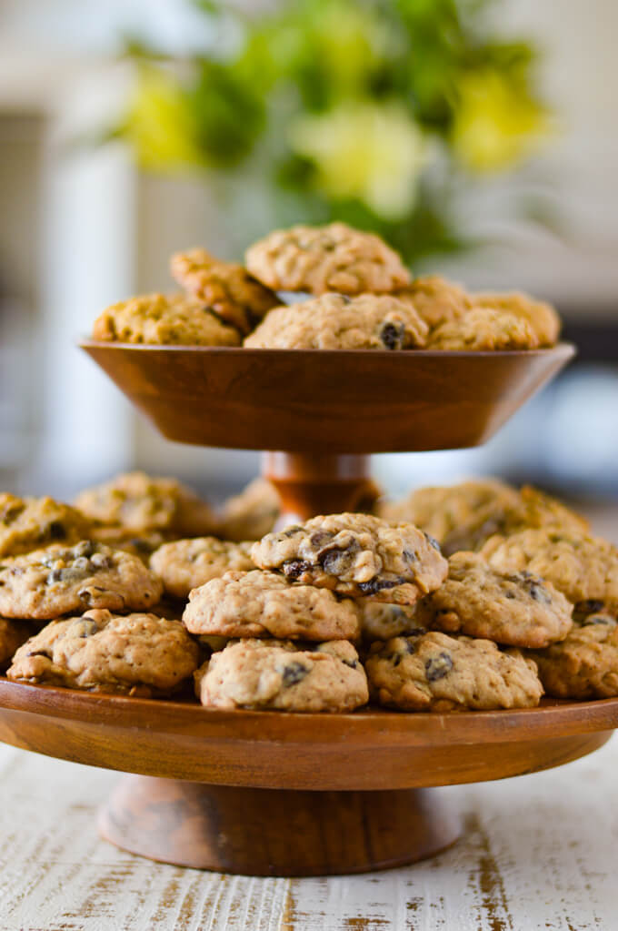 A tiered wooden platter full of oatmeal raisin cookies with a bouquet of flowers blurred in the background.