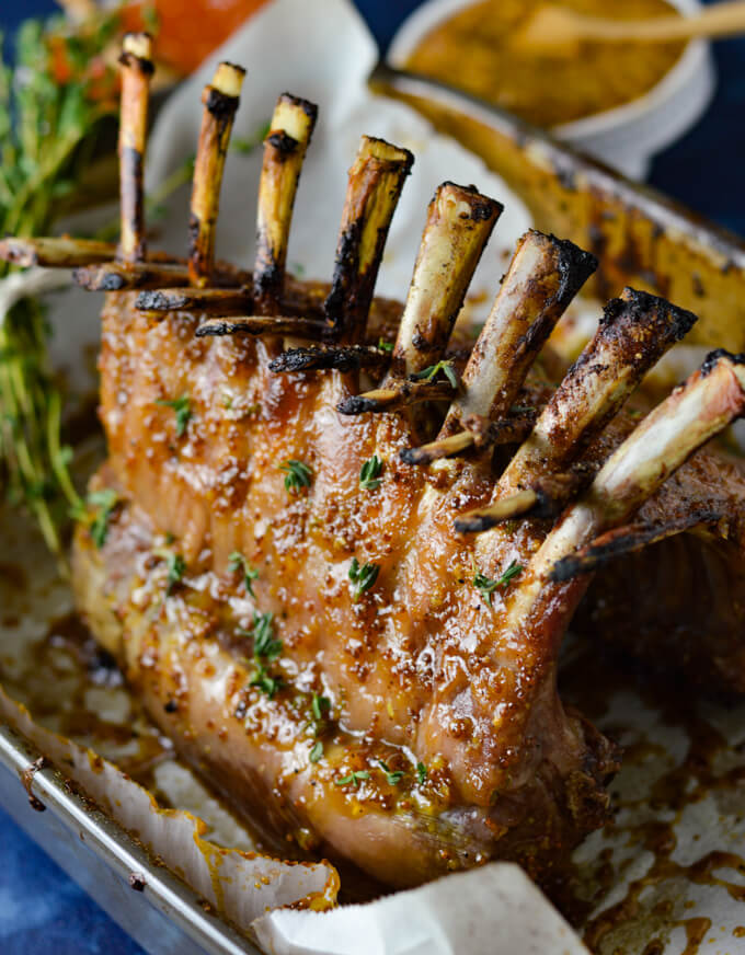 An oven roasted rack of lamb sitting in a shallow pan with a stalk of thyme, bowl of apricot sauce and bowl of mustard in the background.