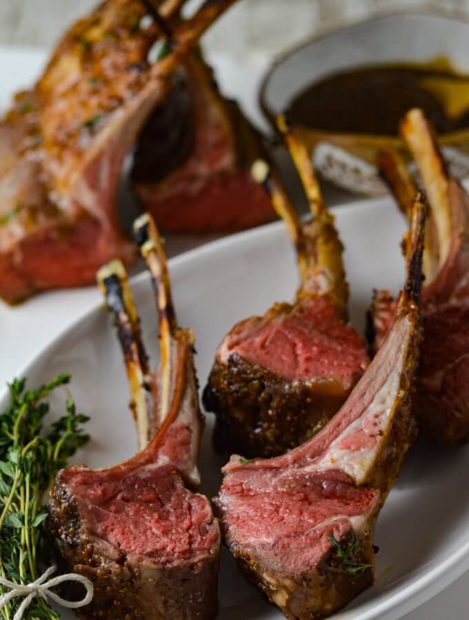 Four lamb chops cooked rare sitting on a white plate with a bundle of thyme next to it. Two partial racks of lambs are in the background with a bowl of apricot mustard sauce.