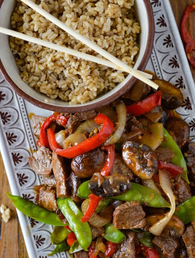 A serving platter of flank steak stir fry next to a bowl of brown rice and wooden chop sticks.