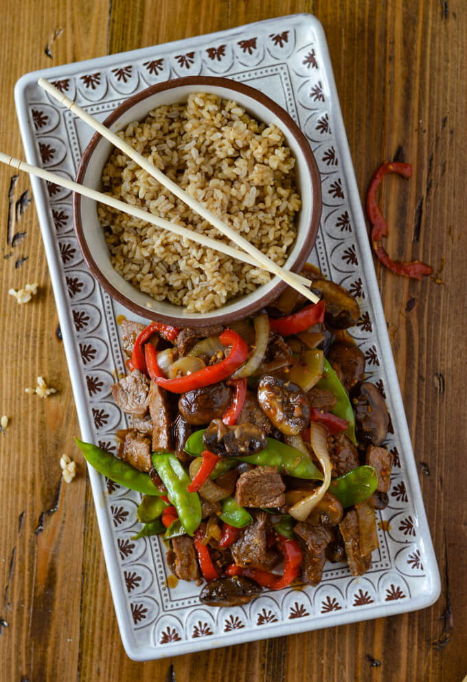 A serving platter of flank steak stir fry next to a bowl of brown rice and wooden chop sticks. 