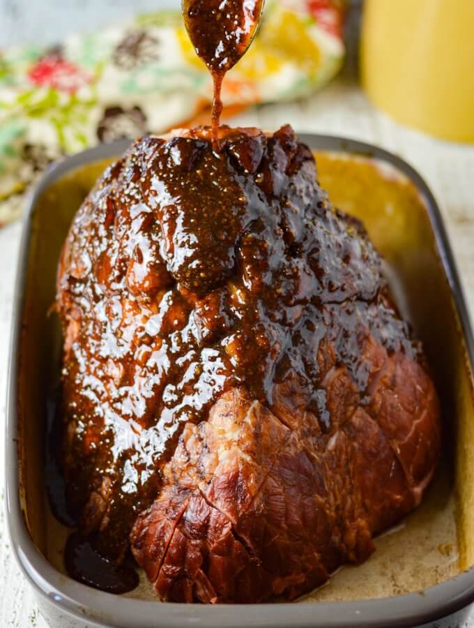 A bone-in ham with brown sugar glaze being poured over it from a spoon above. It sits in a shallow roasting pan ready to go in the oven.