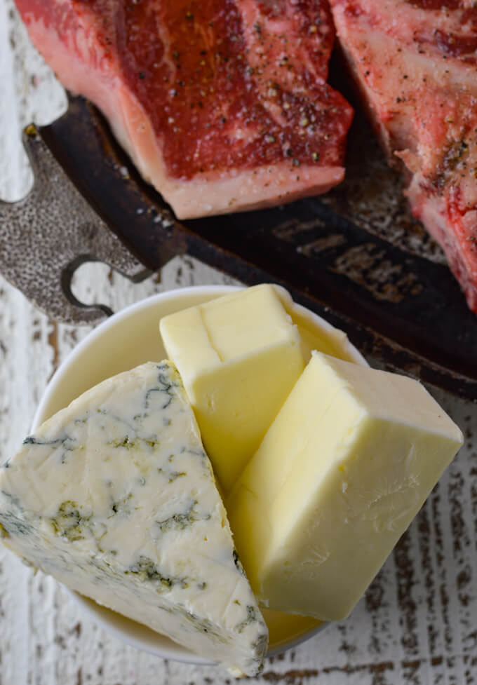 A small white bowl holding a chunck of blue cheese and 2 half sticks of butter. This sits next to a pan where you can see a partial t-bone steak.
