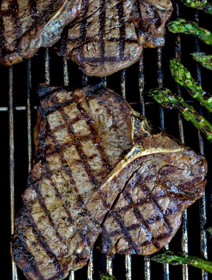 Two cooked t-bone steaks are on the grill with crisscross grill marks on them. Asparagus sits next to them on the grill.