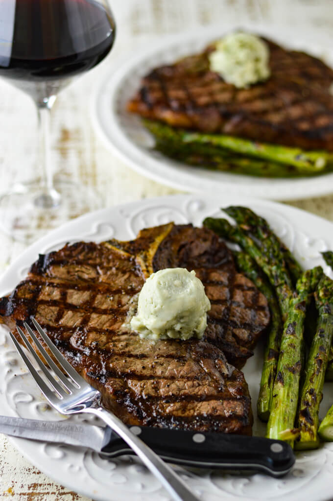 A grilled t-bone steak on a white plate with a round of blue-cheese butter melting on top. A fork and a steak knife sit in front and grilled asparagus is on the plate next to the steak. A another plate with a t-bone steak and asparagus sits in the background. A glass of red wine is off to the side of the photo.