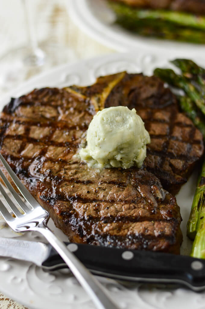 A grilled t-bone steak on a white plate with a round of blue-cheese butter melting on top. A fork and a steak knife sit in front and grilled asparagus is on the plate next to the steak.