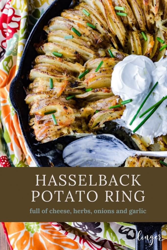 A hasselback potato ring in a cast iron skillet garnished with three dollops of sour cream and chopped chives. A colorful napkin sits beside the skillet. A spoonful has been taken out and the spoon with sour cream is still in the skillet.