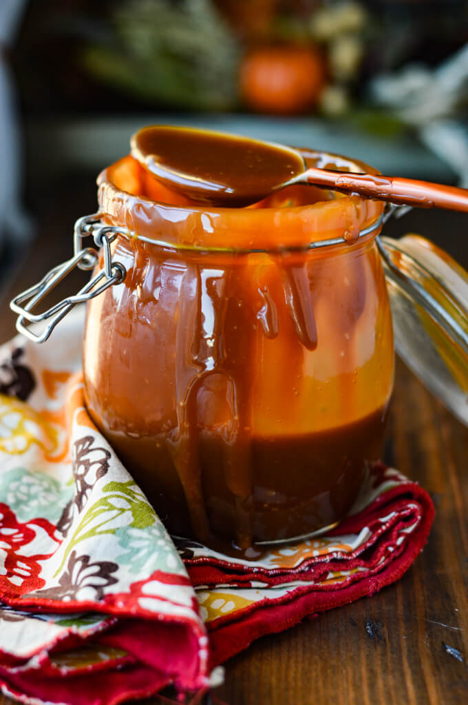 A messy jar of homemade caramel sauce sits on a colorful napkin with sauce dripping from the jar. A spoon of sauce sits on top the jar. 