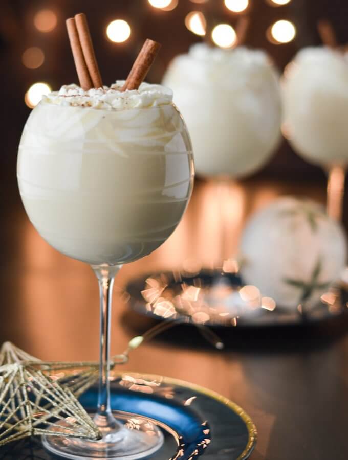 Three glasses of homemade eggnog topped with whipped cream and cinnamon sticks. A few glass ornaments sit next to them. Blurred Christmas lights are in the background.