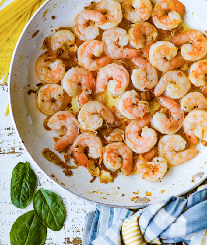 A skillet with cooked shrimp. Dry pasta, a few spinach leaves and a blue napkin lays around the skillet.