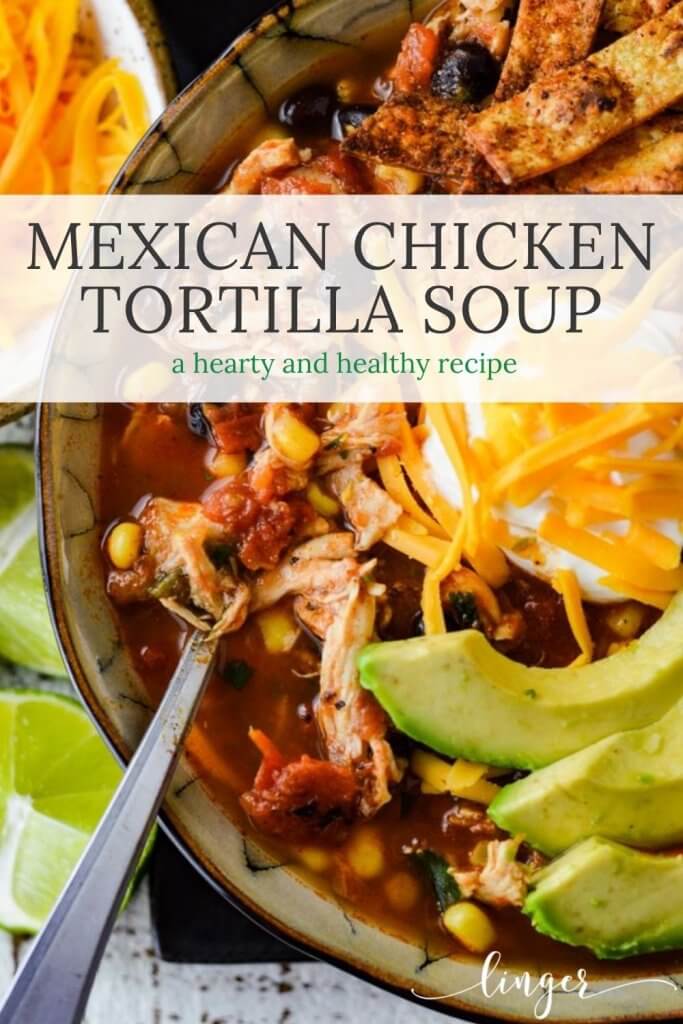 A close-up photo of a bowl of homemade chicken tortilla soup with tortilla strips, slices of avocado, a dollop of sour cream and shredded cheese. A small bowl of shredded cheese and some cut up limes sit next to the bowl of soup.