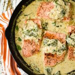 Four pan-seared salmon fillets sit in a creamy mustard and spinach sauce in a cast-iron skillet. A orange and white striped towel sits beside the pan.