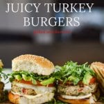 Two turkey burgers sitting on a wooden tray. Layered with cheese, tomato, avocado and arugula. One top bun with grill marks leans next to one of the burgers.