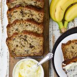 Slices of banana nut bread sit on a wooden cutting board with a small bowl of butter. Two slices of bread, one buttered and three bananas sit next to it.