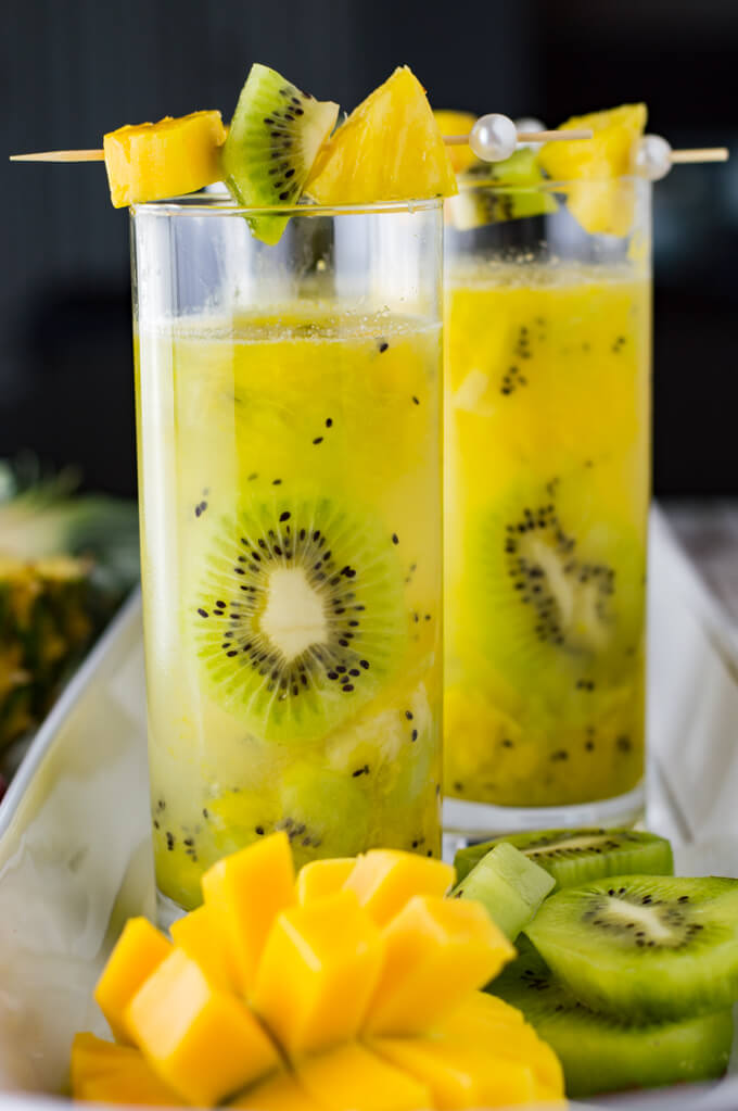 Two glasses of tropical cocktails garnished with pineapple, kiwi and mango sit on a white serving tray. Fresh fruit sit next to the glasses.