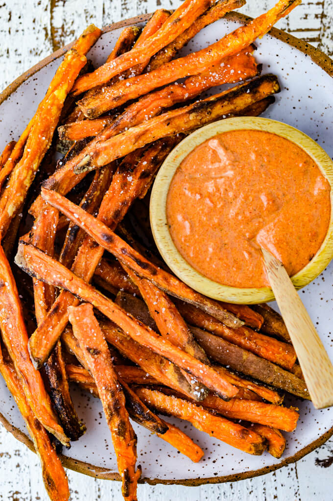 A bowl of Spicy Roasted Tomato and Garlic Aioli with a small wooden spoon in it. Sweet potato fries are on the plate next to the bowl.