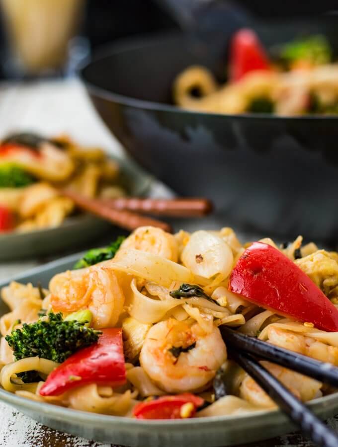 A front view of a bowl of Thai Drunken Noodles with black chopsticks. Another bowl and a wok with the same is blurred in the background.