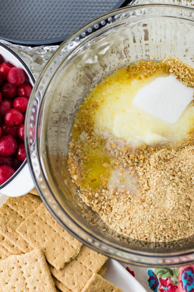 A clear mixing bowl with graham cracker crumbs, sugar and melted butter. A bowl of tart cherries and whole graham crackers sit next to the bowl.