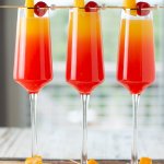 Three tequila sunrise mimosa cocktails with cocktail pics of cherries and orange slices sitting on a wooden board.