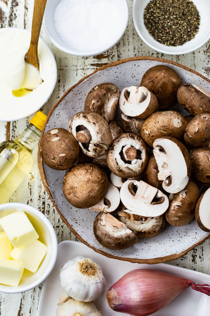 A bowl of raw mushrooms with butter shallots, sliced garlic and salt and pepper sit around the bowl.