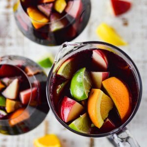 A photo of a pitcher of red wine sangria with chunks of fresh apples, oranges, lemons and limes. Two glasses filled with the sangria sit beside the pitcher. Chucks of the fresh fruit are scattered on the board below.