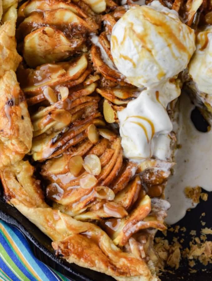 Caramel Apple Galette with a puff pastry crust in a cast iron pan. A blue striped napkin sits next to it. Three scoops of ice cream sit on the apples. A slice is taken out of it and the ice cream is melting into the pan.